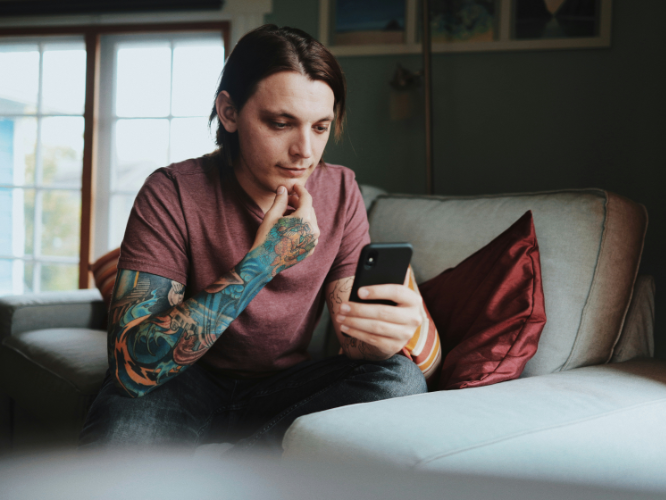 A tattooed man in a maroon t-shirt looking at a smartphone on a sofa. Credit: Charley Pangus, via Unsplash.