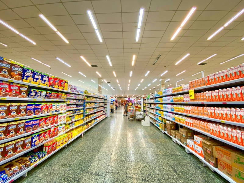 A wide supermarket aisle lined with shelves of packaged groceries, viewed toward the centre of the store. Credit: Nathália Rosa, via Unsplash.