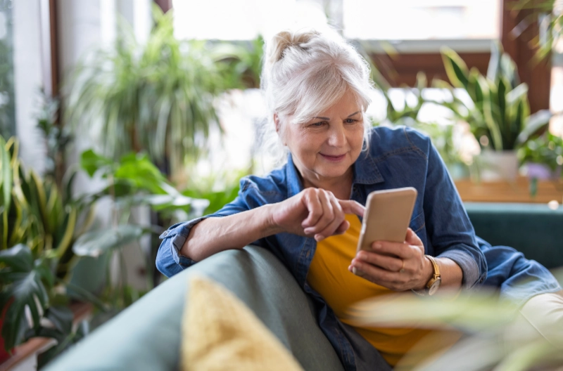 Smiling senior woman using smart phone while sitting on sofa at home. Credit: Piksel, via iStock.